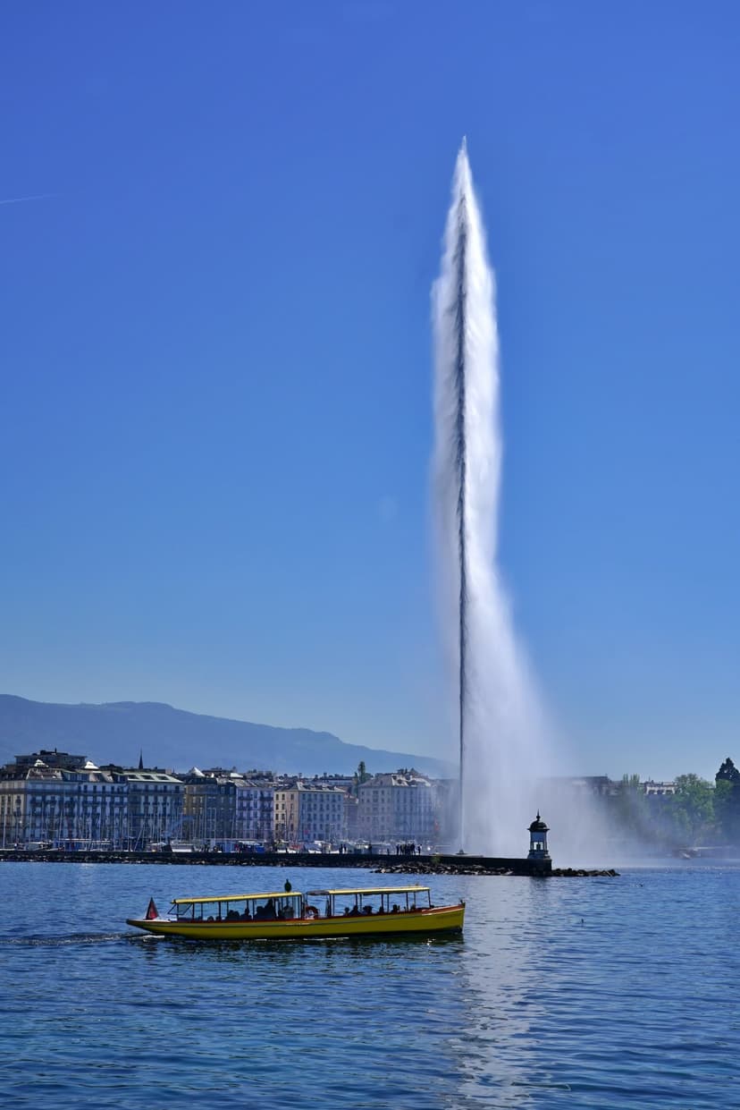 Jet d'Eau fountain spraying water upward beside a yellow boat on Lake Geneva with distant mountains.