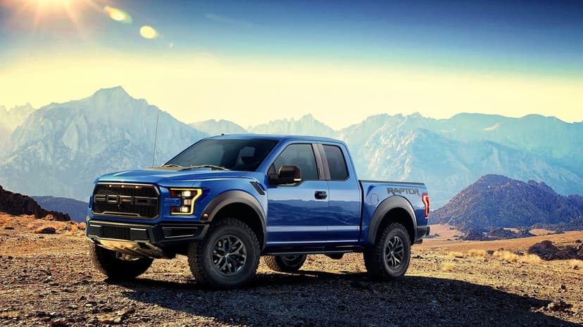 Blue Ford F-150 Raptor truck parked on a rocky desert floor with mountains and sun flare in the background.