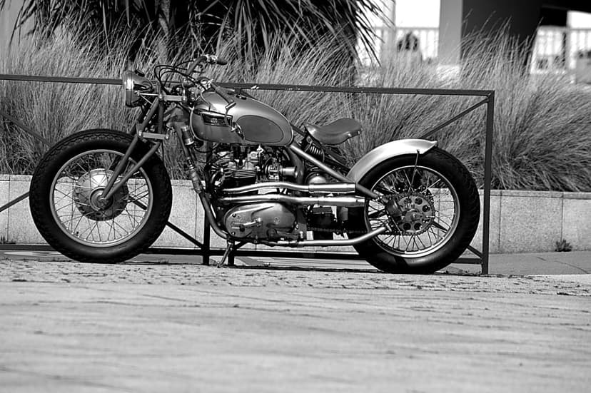 Custom motorcycle with chrome exhaust and spoked wheels parked on a paved ground in a black and white portrait.