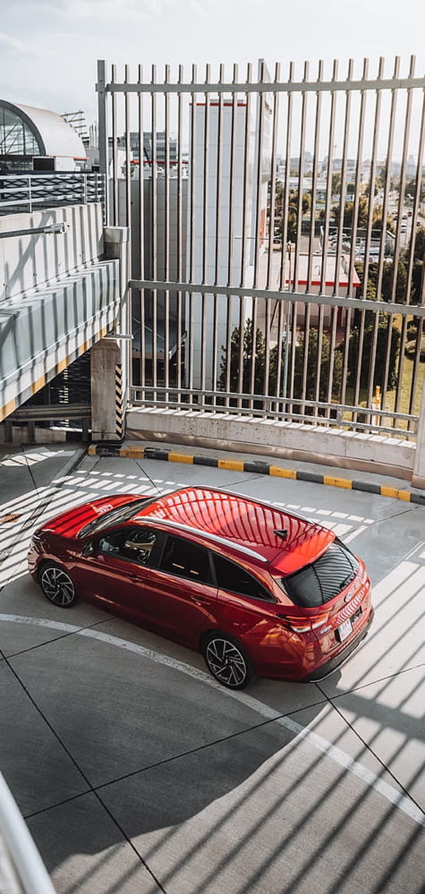 Red station wagon parked on asphalt with long striped shadows from a fence in a concrete urban parking garage.