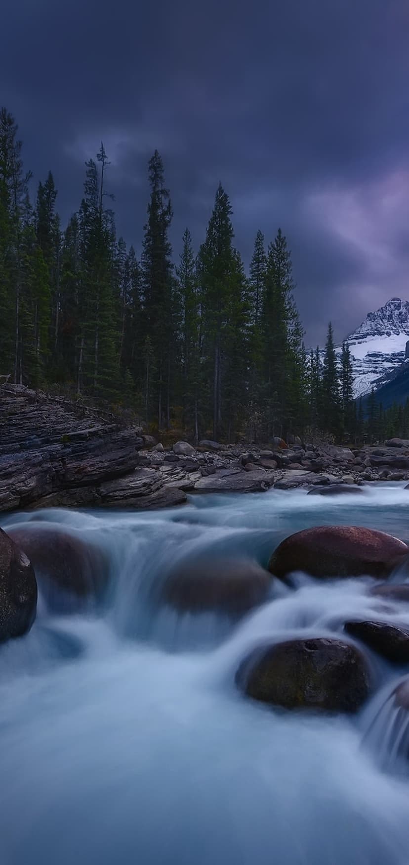 Cascading river water flows over mossy boulders with a snow-capped mountain peak under a dark twilight sky.