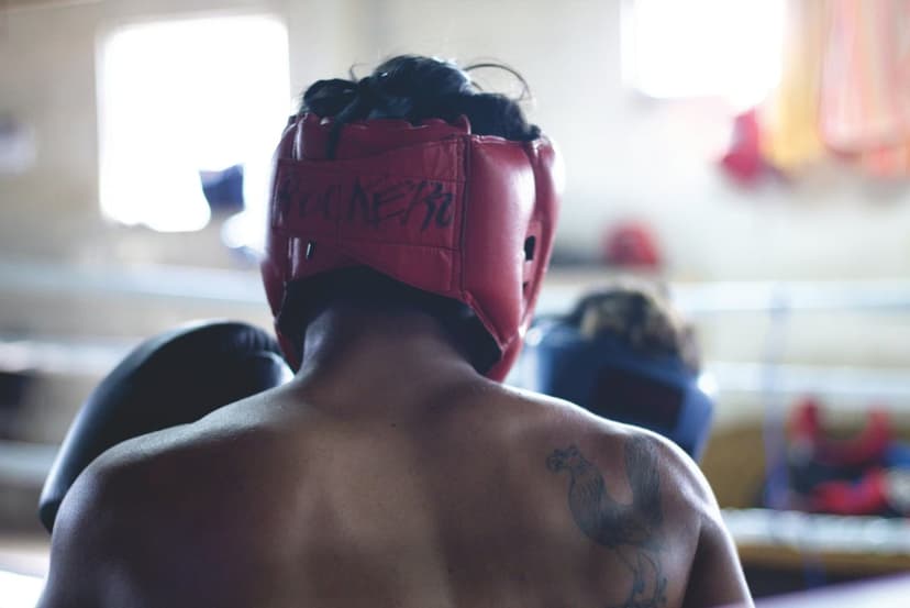 Boxer from behind wearing red protective headgear and black gloves with a shoulder tattoo in a boxing ring.