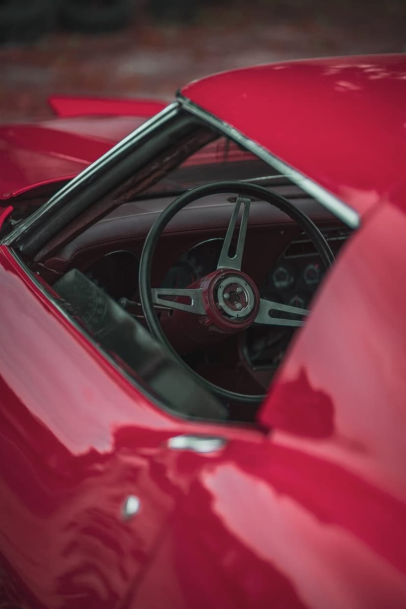 Vintage steering wheel with chrome spokes and a burgundy center hub inside a classic red sports car cabin.