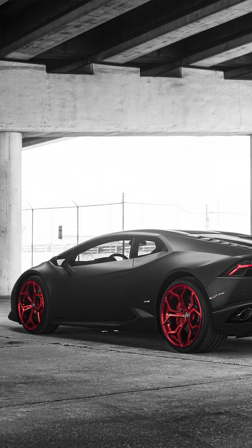 Matte black Lamborghini with bright red wheels parked on asphalt beneath a massive grey concrete bridge pillar.
