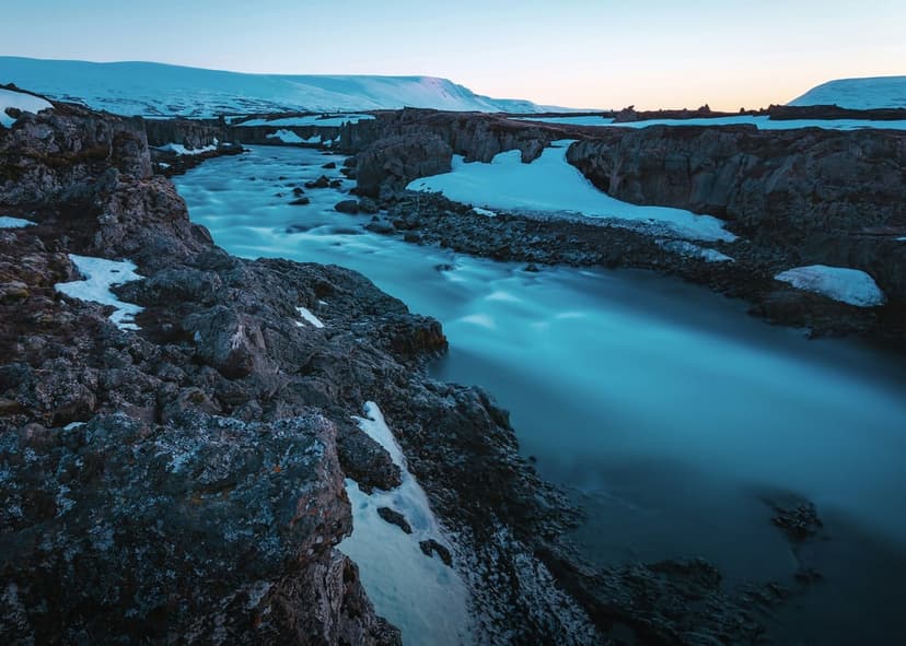 Blue river water flows between snow-covered volcanic rocks under soft light in a vertical Icelandic landscape.