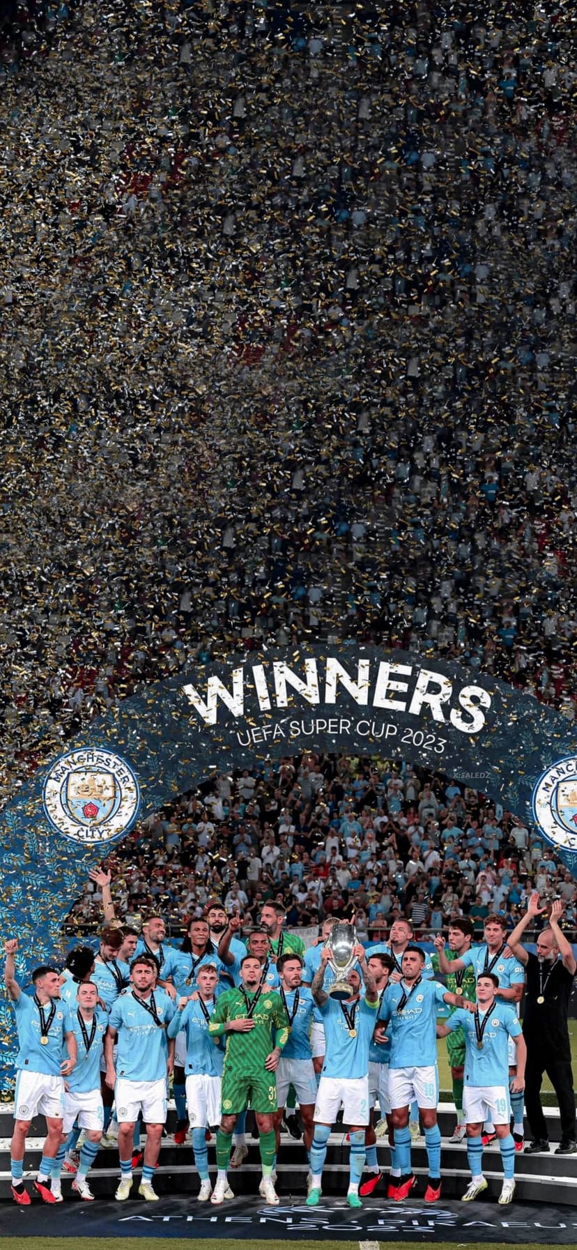 Manchester City football players raise a silver trophy during a night celebration with blue and white confetti.
