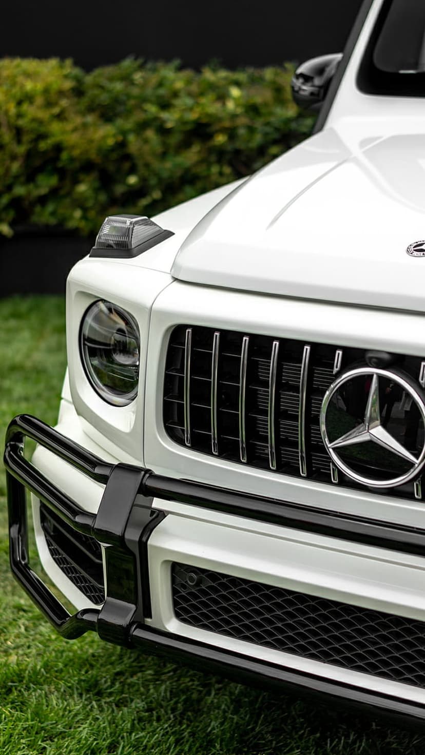 Mercedes-Benz G-Class grille with chrome star emblem, round headlight, and black bumper over green grass.