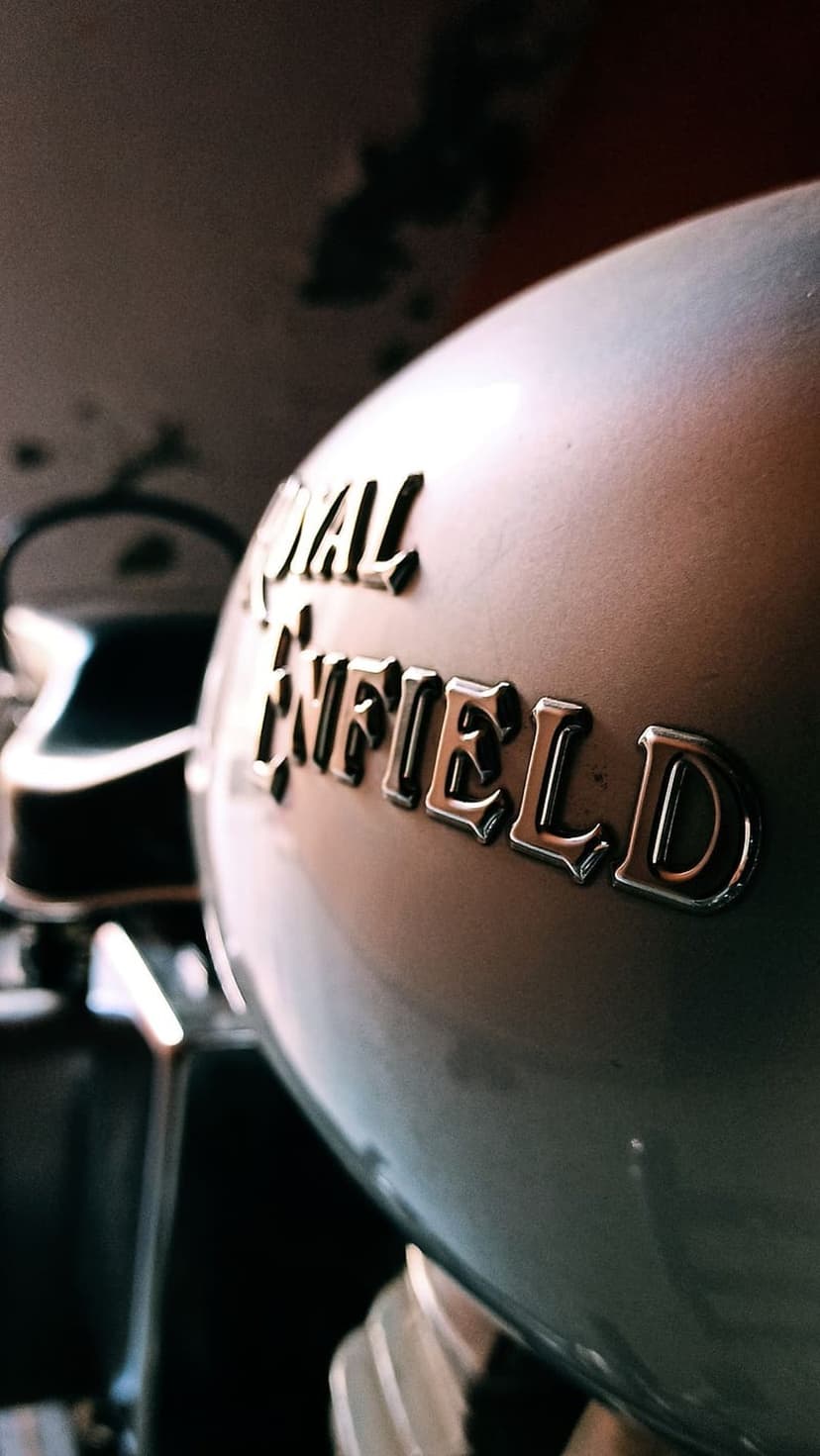 Royal Enfield chrome emblem on a metallic silver motorcycle tank with blurred mechanical parts in the background.