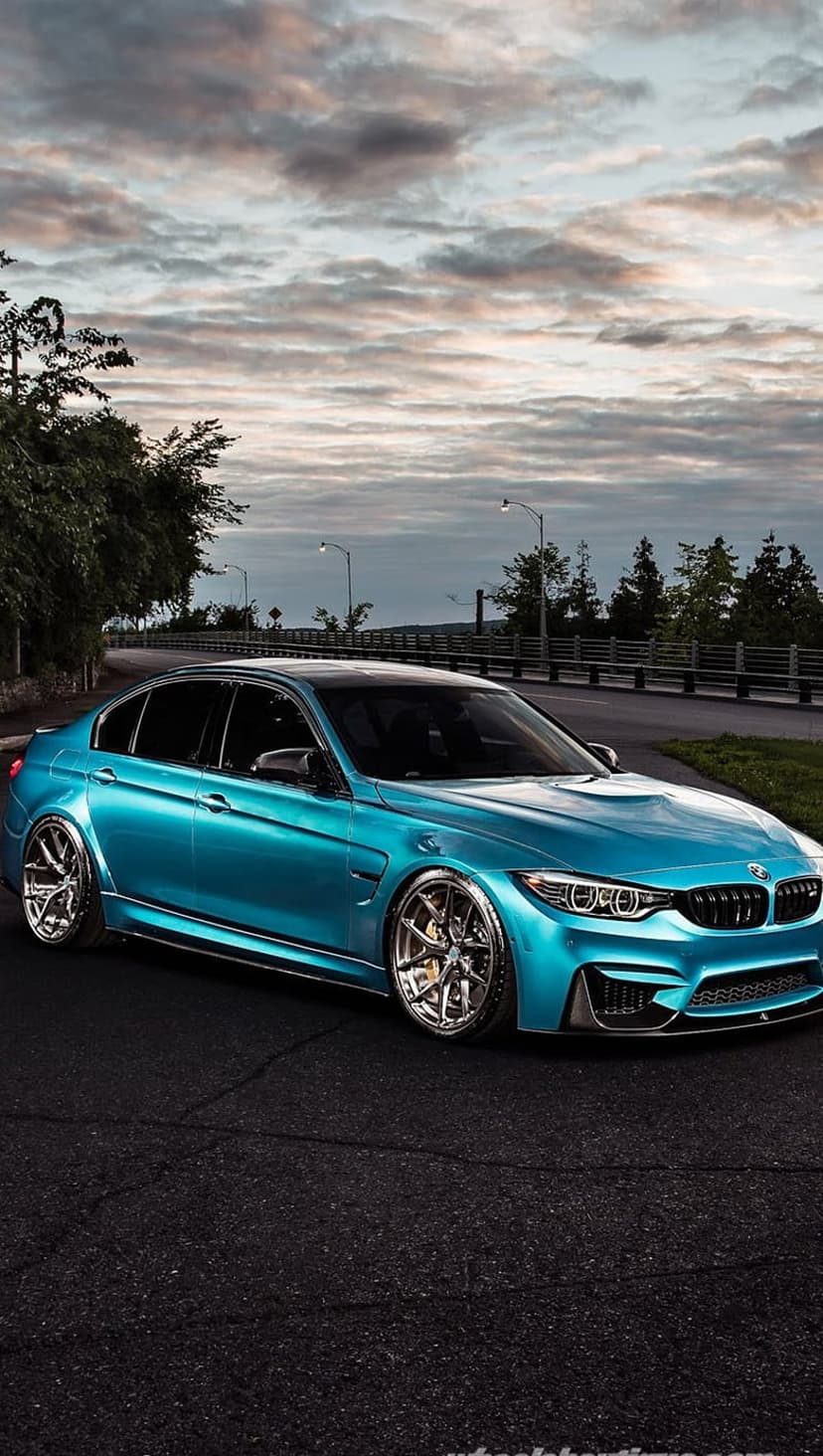 Teal BMW M3 sedan with chrome rims parked on asphalt under a stormy sky with lush forest greenery in the distance.