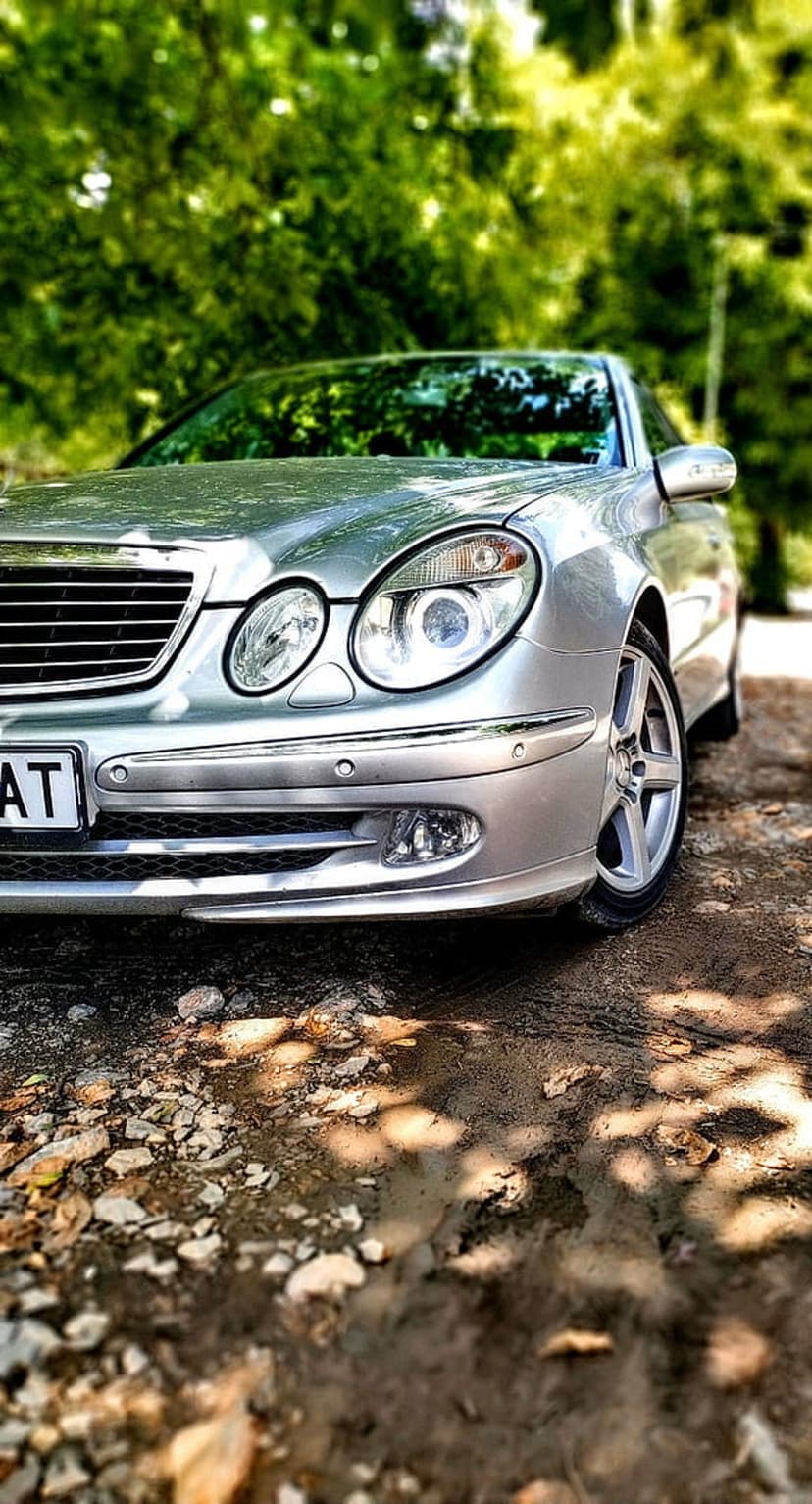 Silver Mercedes E-Class sedan parked on gravel under green trees with dappled sunlight hitting the front grille.