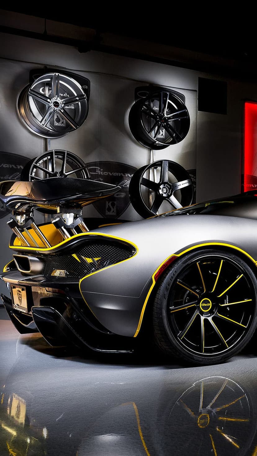 McLaren P1 hypercar with silver paint and yellow trim sits in a showroom before a wall of luxury wheels.