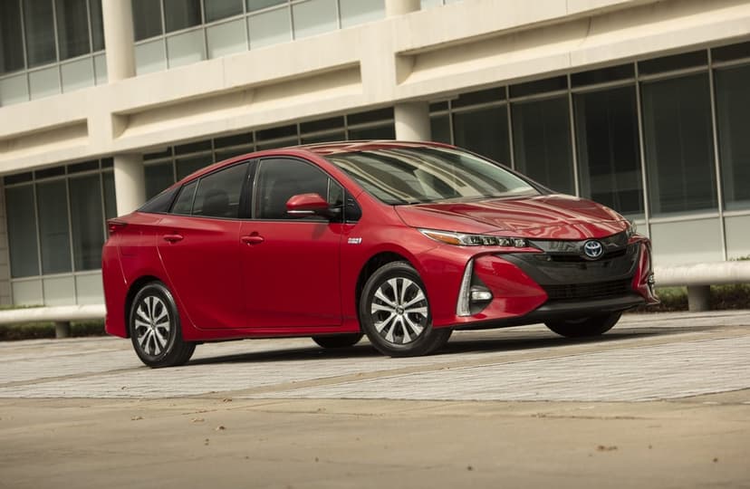 Red Toyota Prius Prime plug-in hybrid sedan parked on concrete in front of a modern glass building.