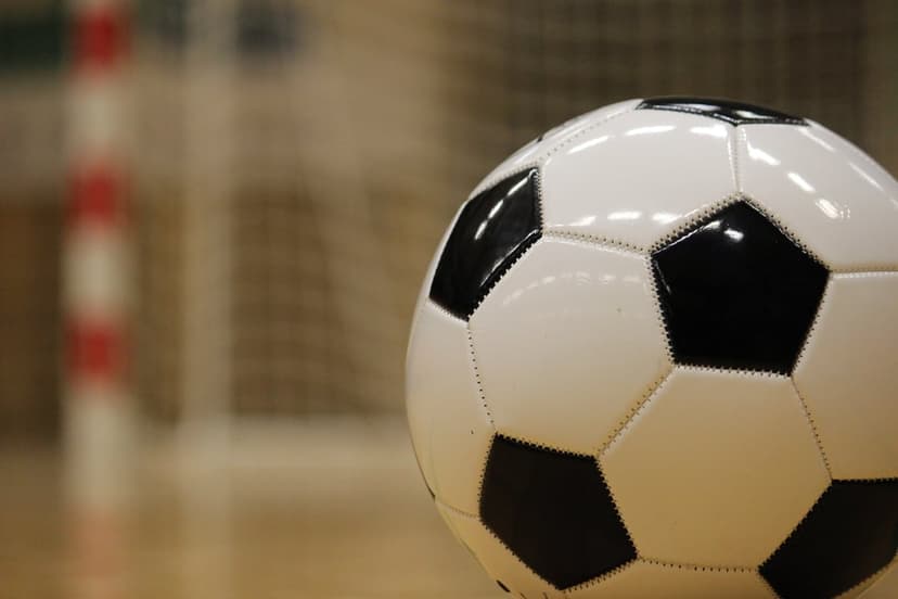 Black and white soccer ball on a wooden floor with a blurred goal net and red striped post in the background.