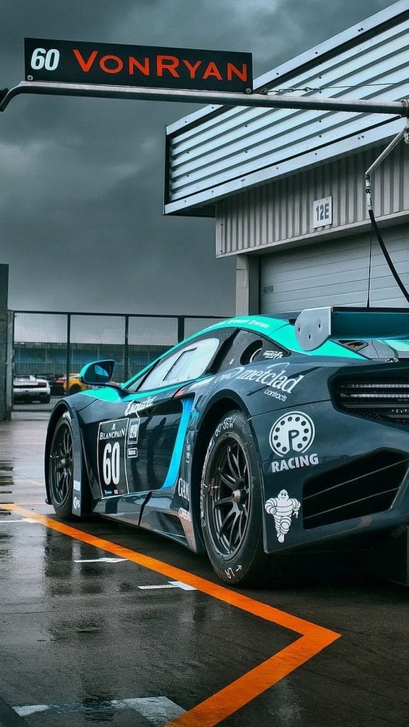 Teal McLaren GT3 race car parked on a wet track with orange reflections under a dark, moody sky.