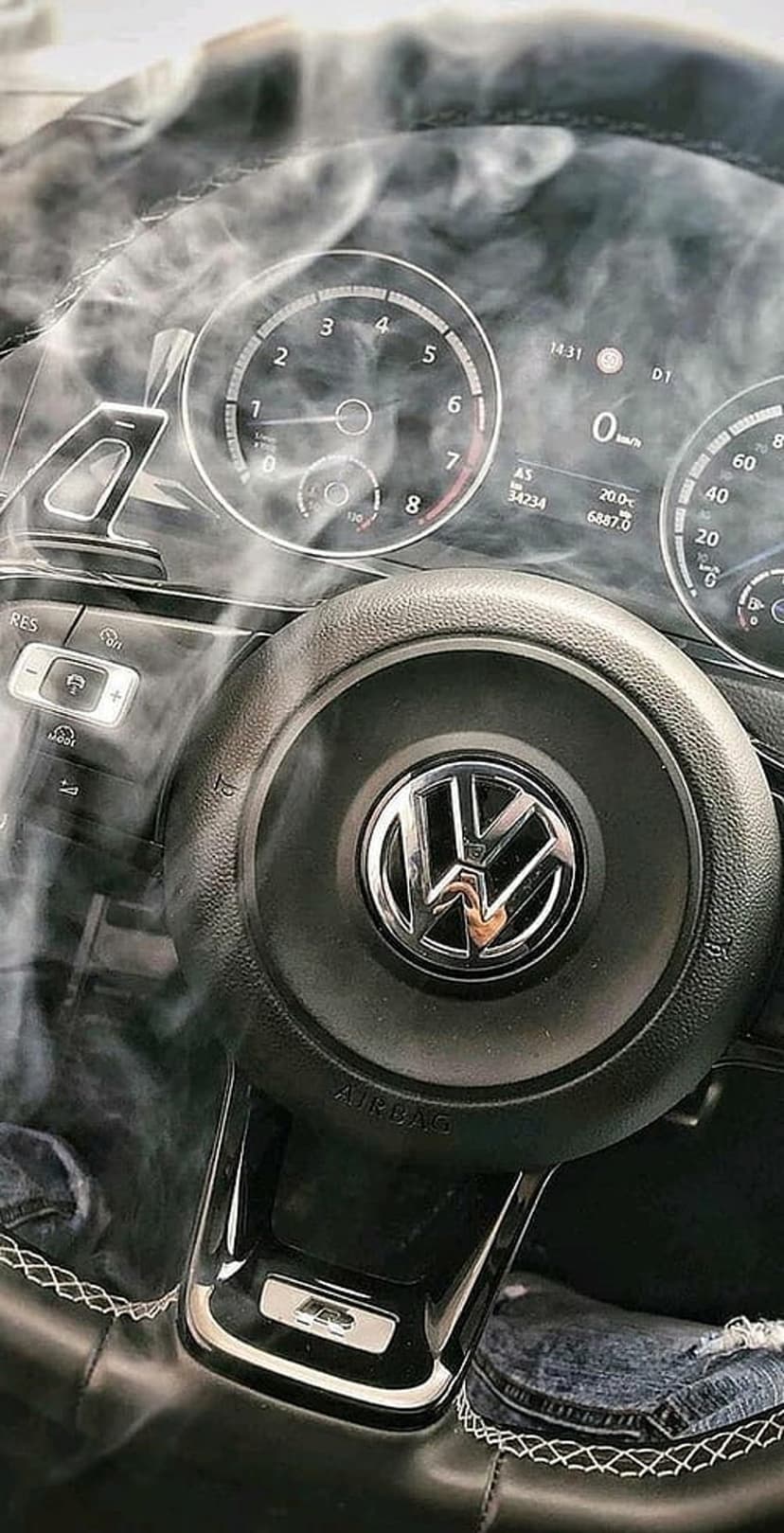 Volkswagen steering wheel and glowing digital dashboard obscured by thin wisps of white smoke in a dark cabin.