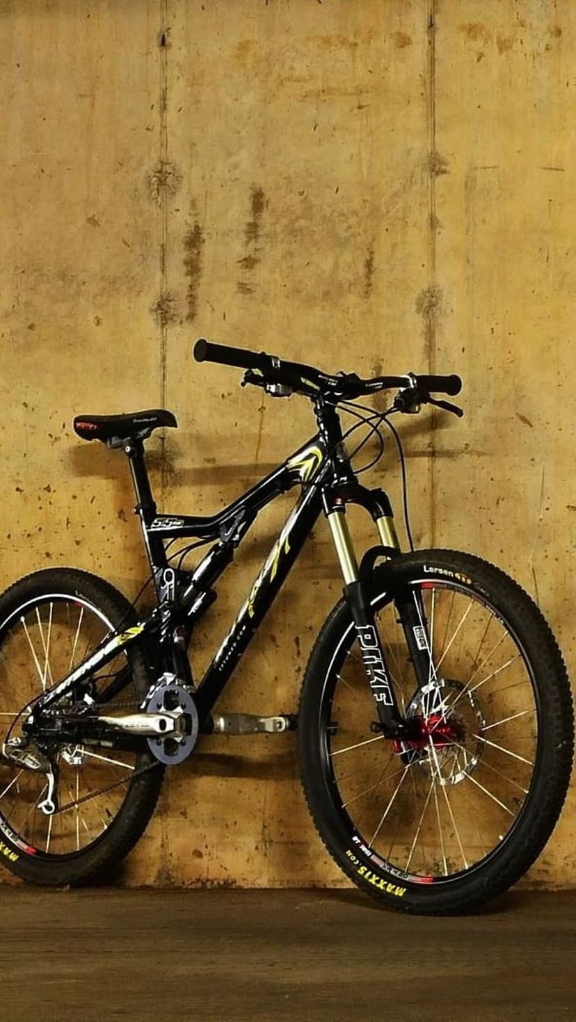Black mountain bike with yellow accents and Maxxis tires leaning against a grey textured concrete wall.