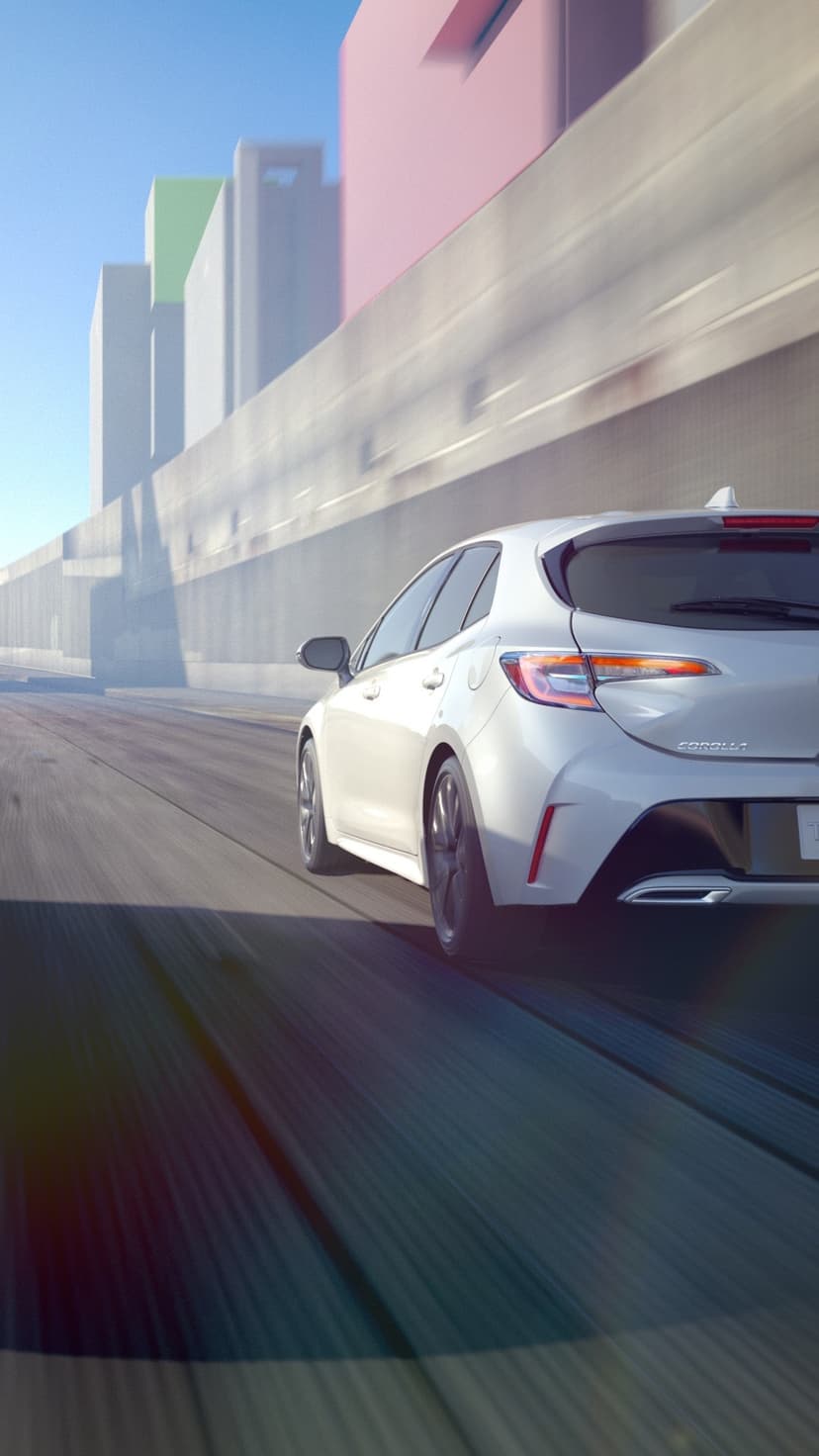 Rear view of a white hatchback car driving fast past pastel buildings with motion blur on the road.