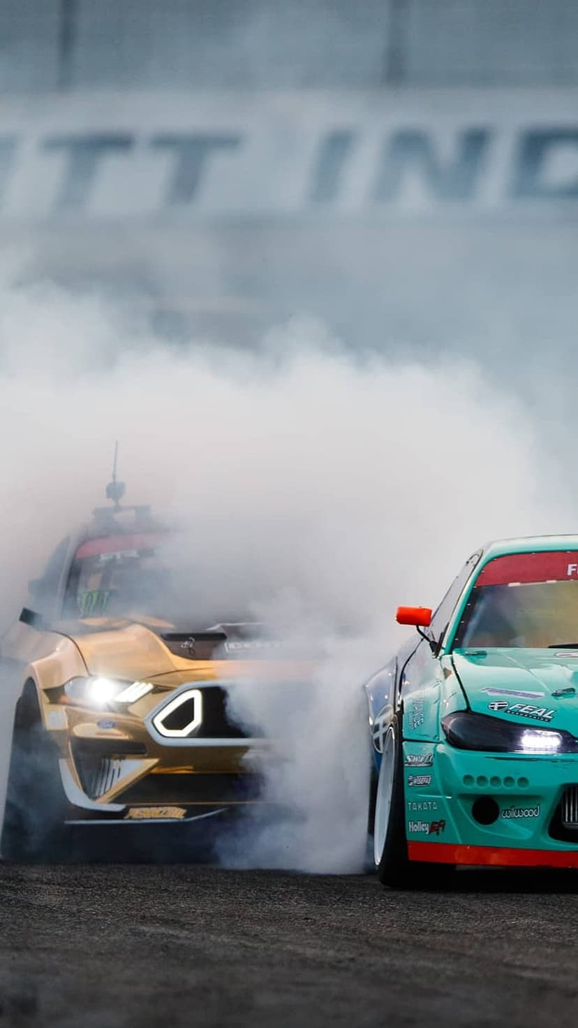 Gold and teal race cars drift on a track, creating thick clouds of white tire smoke against dark asphalt.