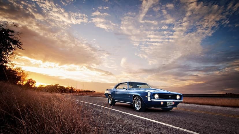 Blue 1969 Chevrolet Camaro SS on a rural road under a dramatic orange and purple sunset sky.
