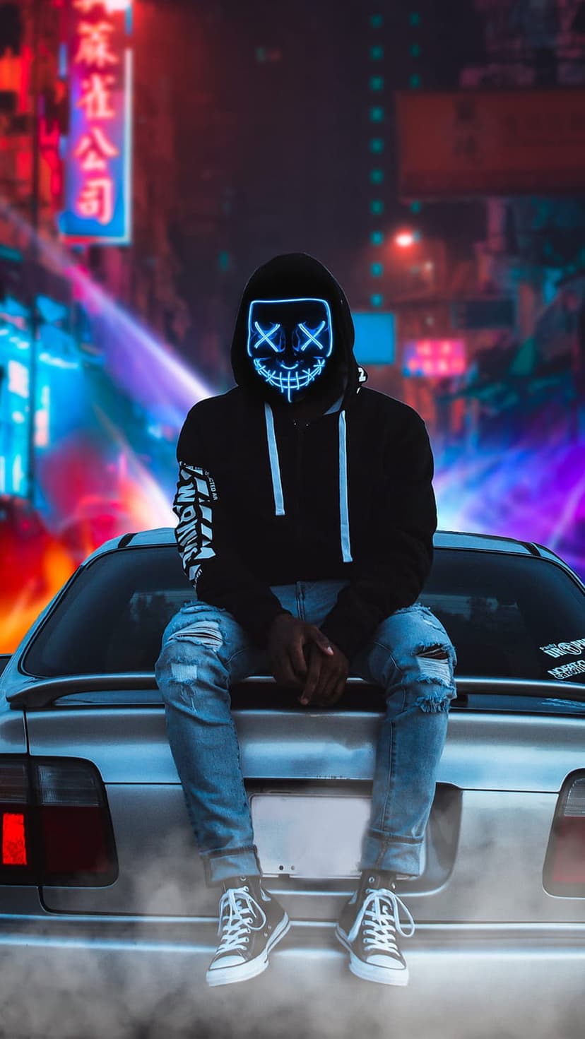 Masked person in a hoodie sits on a car trunk against a vibrant neon cityscape at night with glowing blue lights.