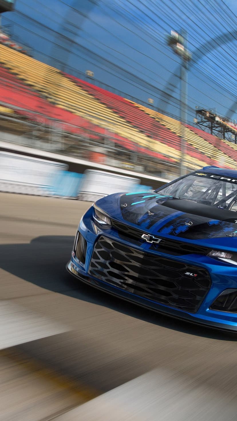 Blue Chevrolet Camaro ZL1 race car speeds on a track past blurred red and yellow grandstands under a blue sky.