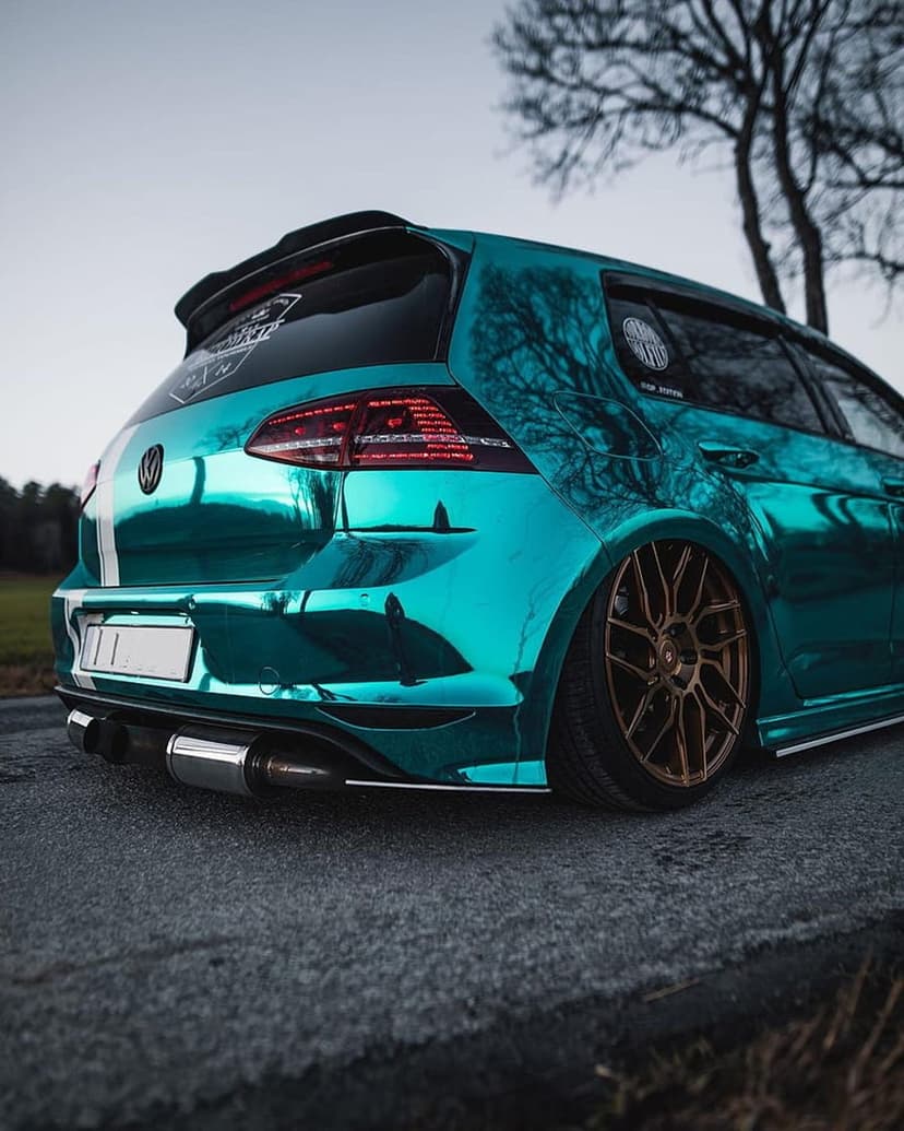 Cyan chrome VW Golf R displays bronze wheels and a custom lowered stance against a reflection of trees.