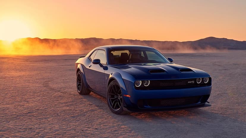 Dark blue Dodge Challenger muscle car on a cracked desert floor during a golden sunset with dust clouds.