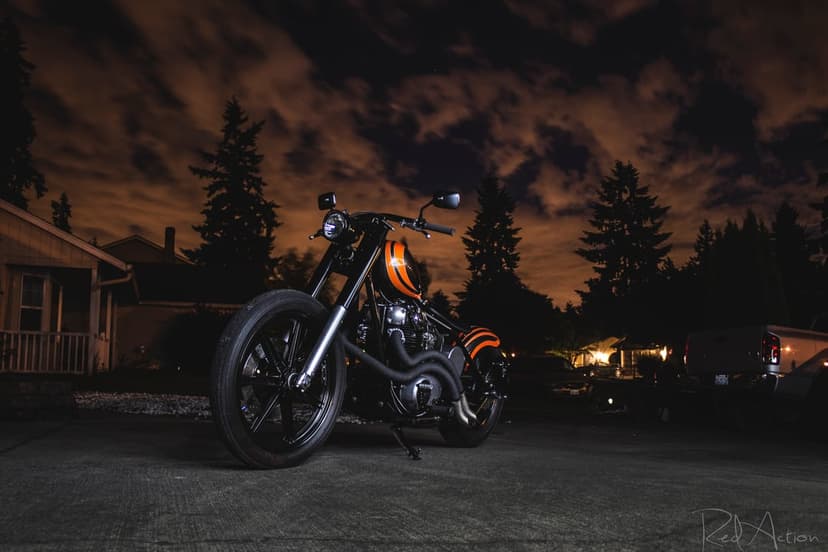 Custom orange chopper motorcycle parked on a dark paved road under a dramatic and illuminated cloudy night sky.