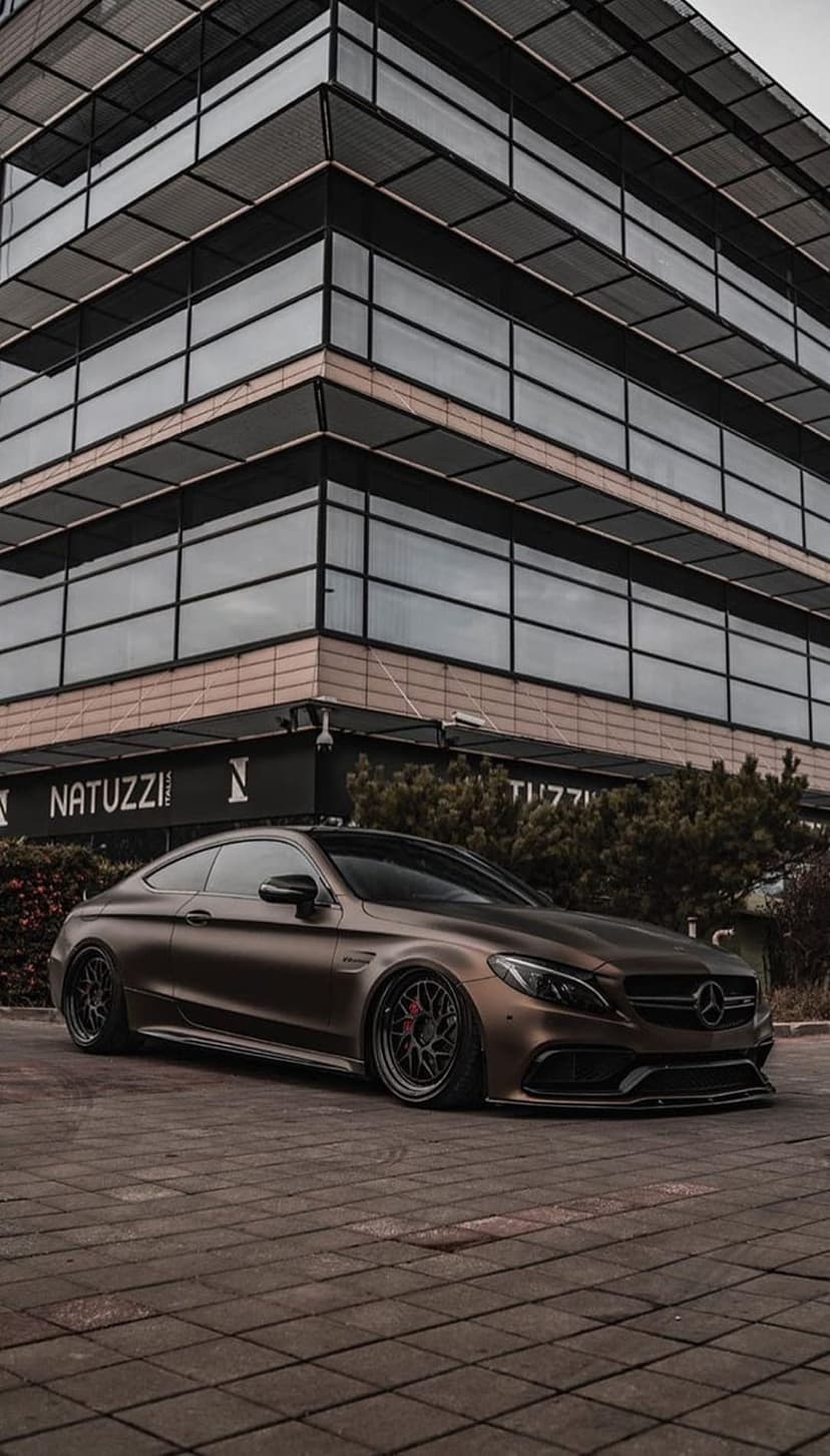 Matte brown Mercedes-AMG C63 Coupe parked on cobblestones in front of a modern glass building with a store sign.