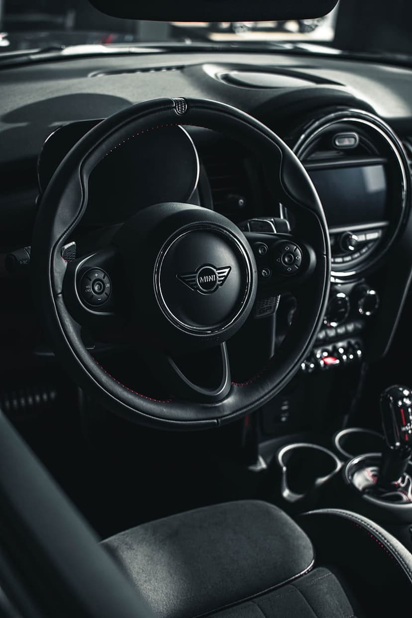 Mini Cooper steering wheel with red stitching and logo rests before a dark dashboard with a central display.