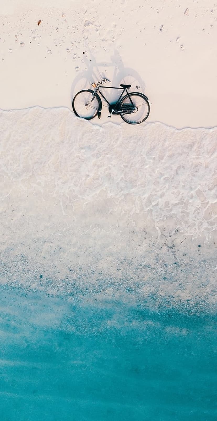 Black vintage bicycle parked on white sand next to turquoise ocean waves from a top-down aerial perspective.