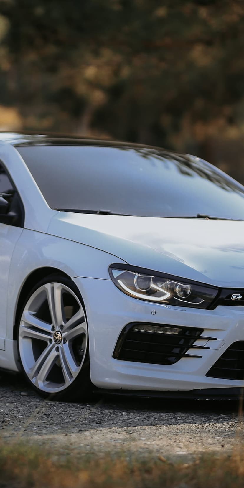 White Volkswagen Scirocco R with LED headlights on a dirt path with blurred autumn trees in the background.