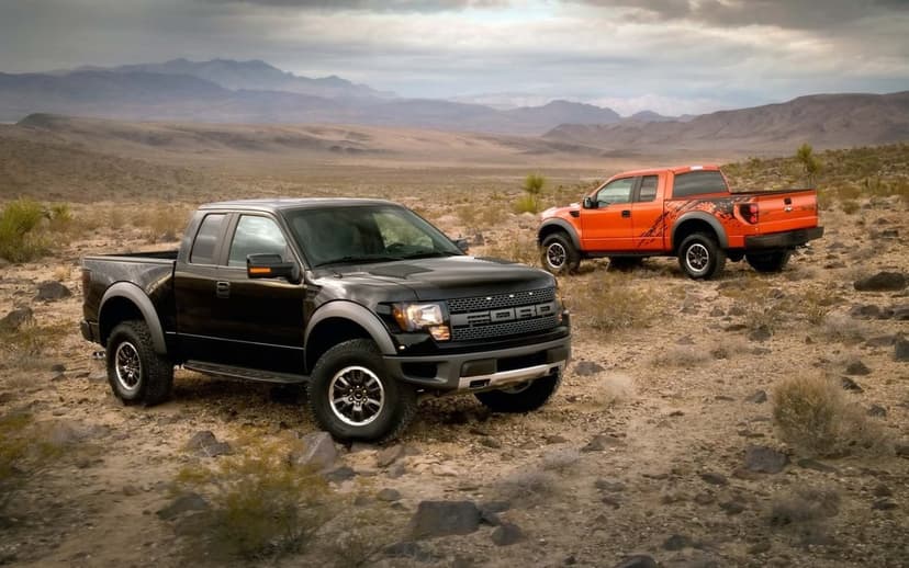 Black and orange Ford Raptor trucks parked on a rocky desert floor with mountains under a cloudy sky.