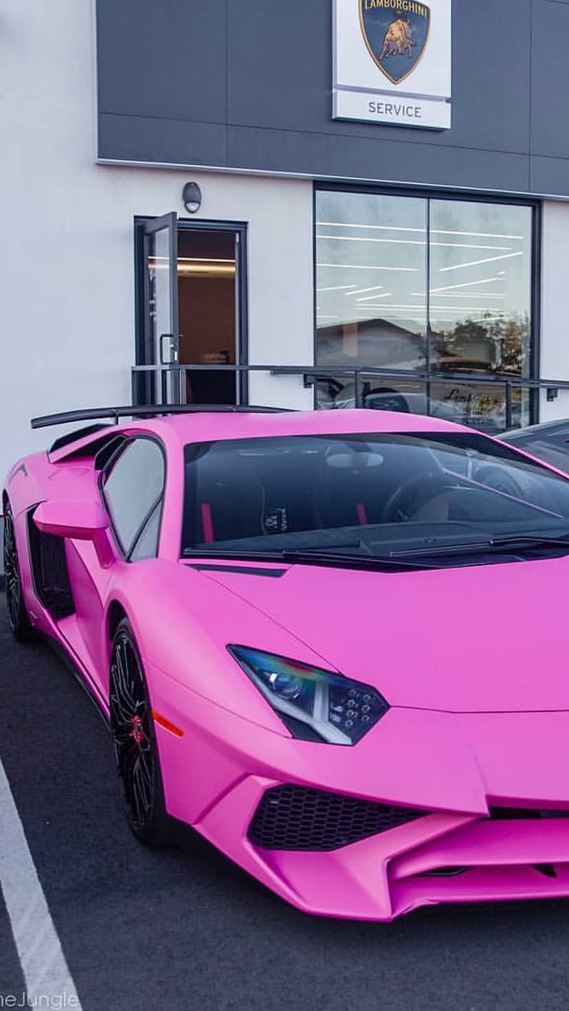 Pink Lamborghini Aventador with a large rear wing parked in front of a modern glass dealership service entrance.