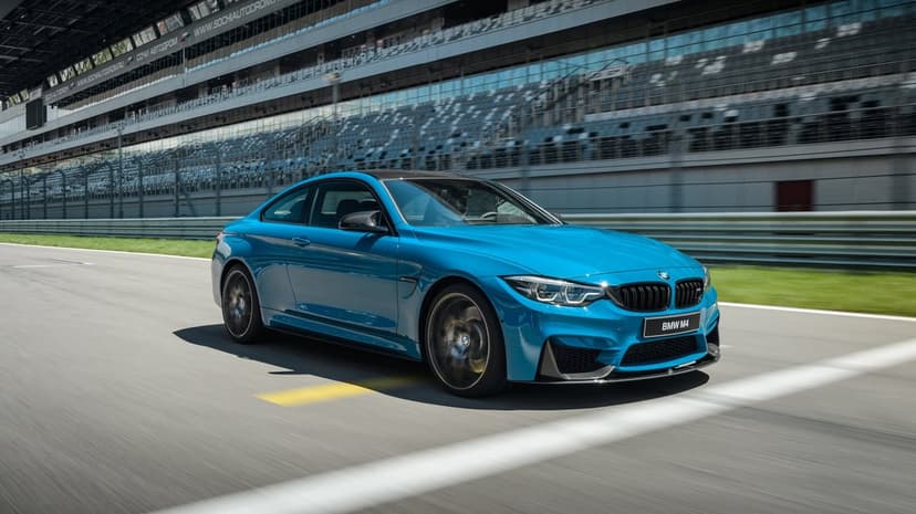 Blue BMW M4 sports car driving fast on a racetrack with motion blur and grandstands in the background.