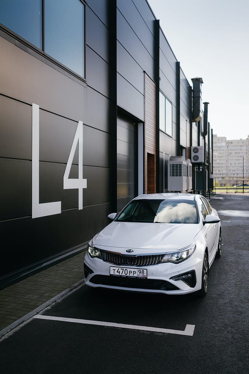 White Kia Optima sedan parked in front of a dark gray contemporary building with a large white L4 graphic.