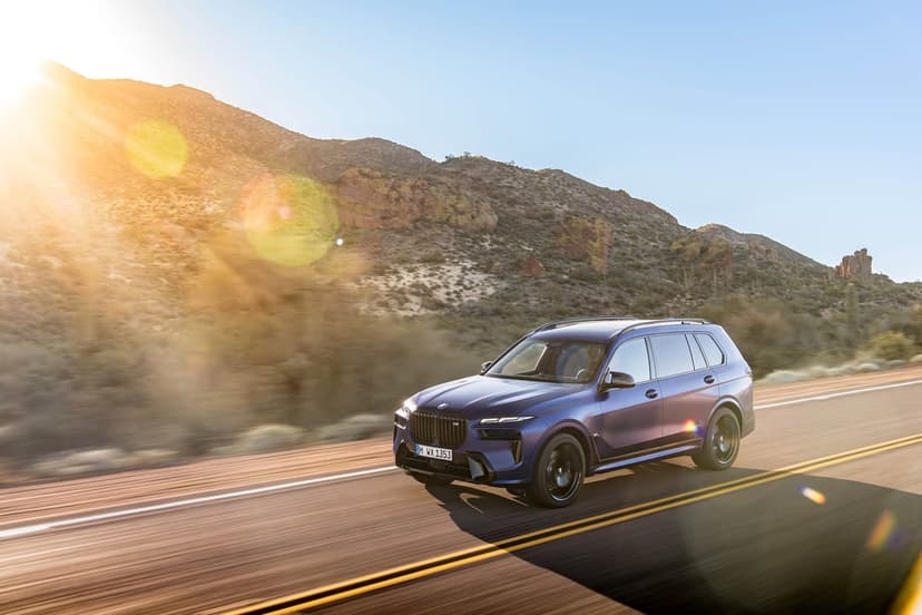 Blue BMW SUV driving on a desert road at sunset with mountains in the background and a bright lens flare.