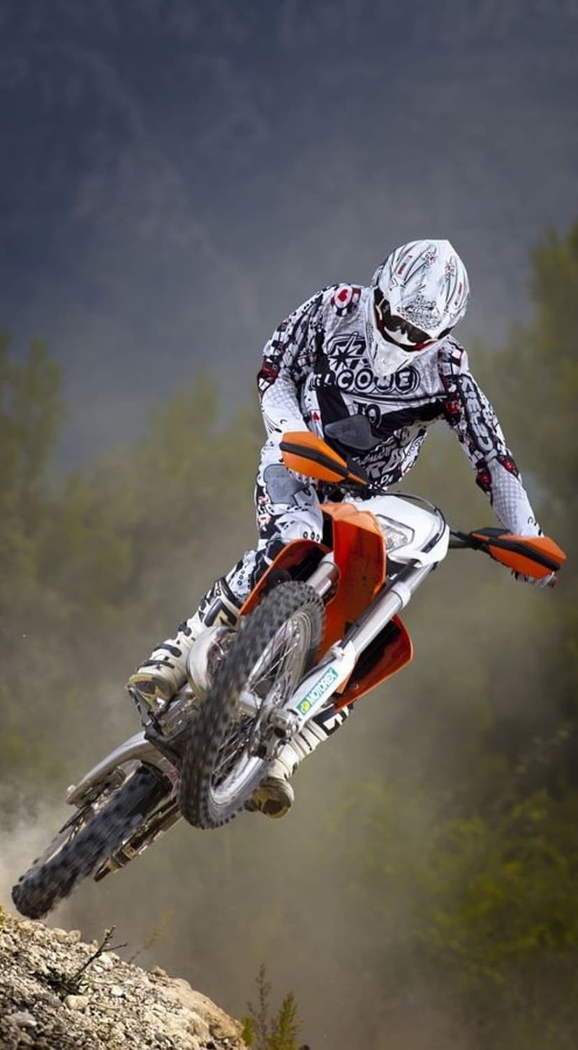 Motocross rider in black and white gear jumps an orange bike over a dusty, rocky course under a hazy sky.