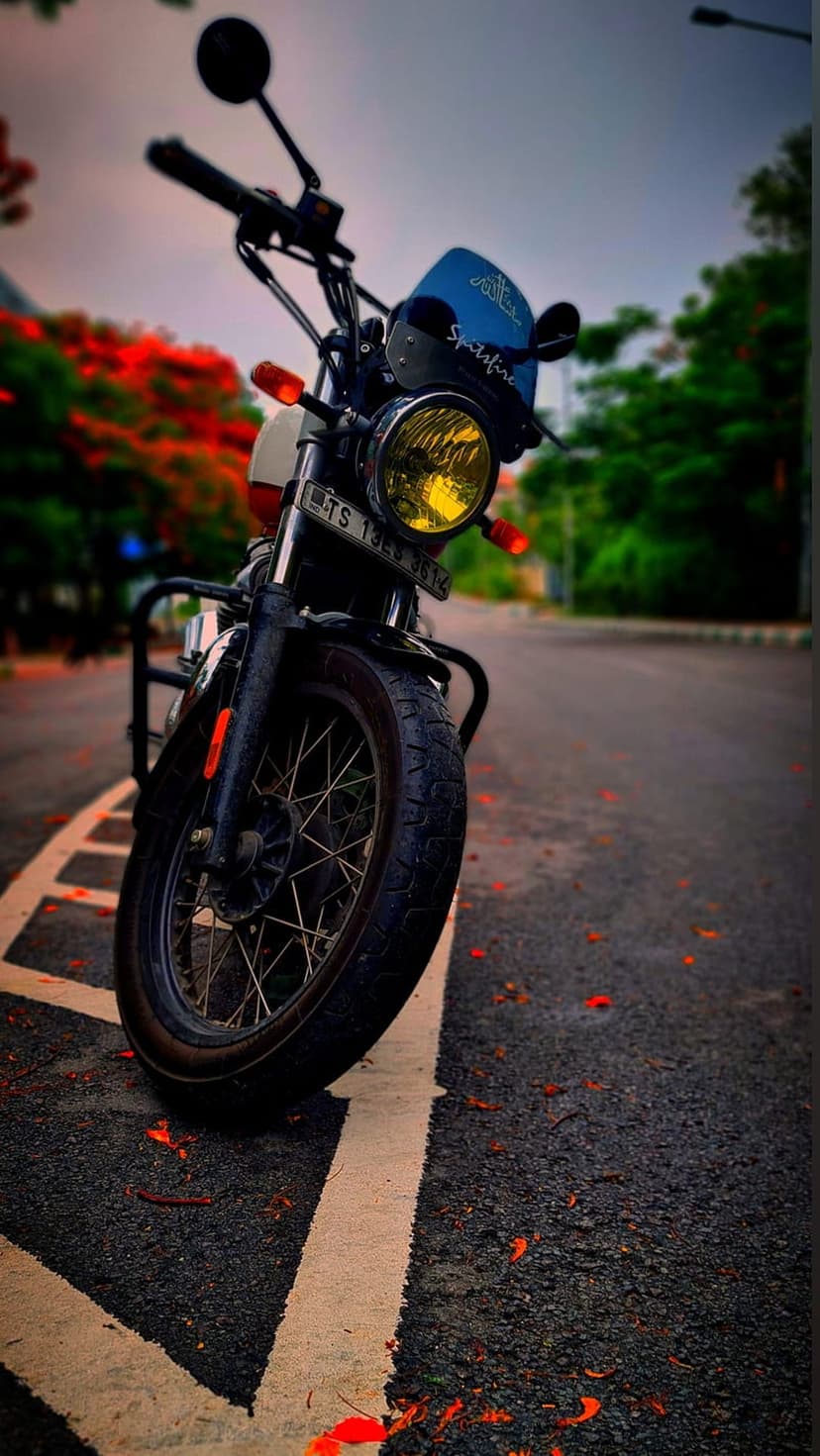 Royal Enfield motorcycle with a yellow headlight and blue windscreen on an asphalt road with orange flowers.