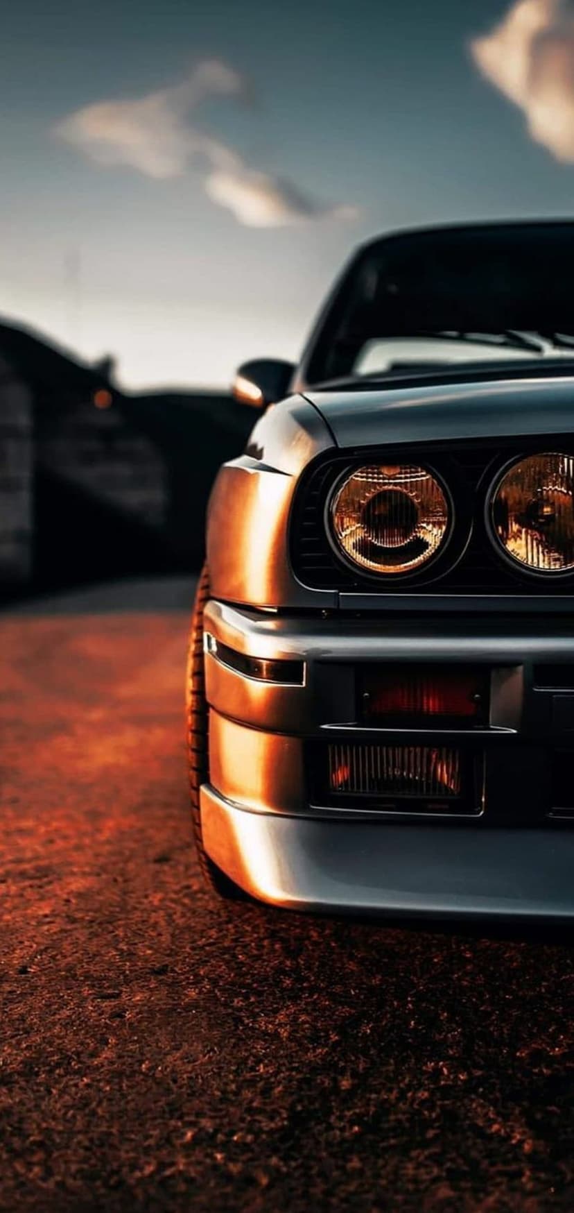 BMW E30 M3 front grill and headlights glowing under a warm sunset with a blurred asphalt road background.