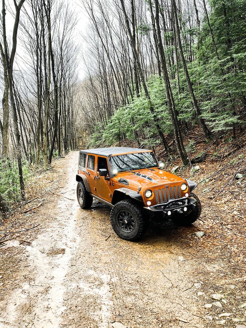 Orange Jeep Wrangler drives through a muddy forest path surrounded by tall trees and damp autumn foliage.