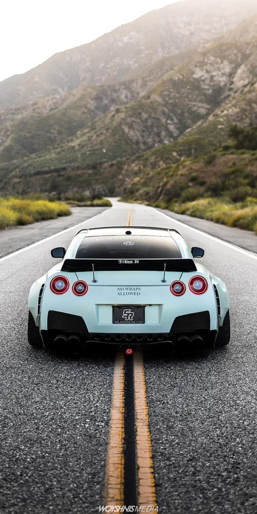 Light blue Nissan GT-R with a widebody kit and rear wing driving on a winding mountain road during a hazy day.