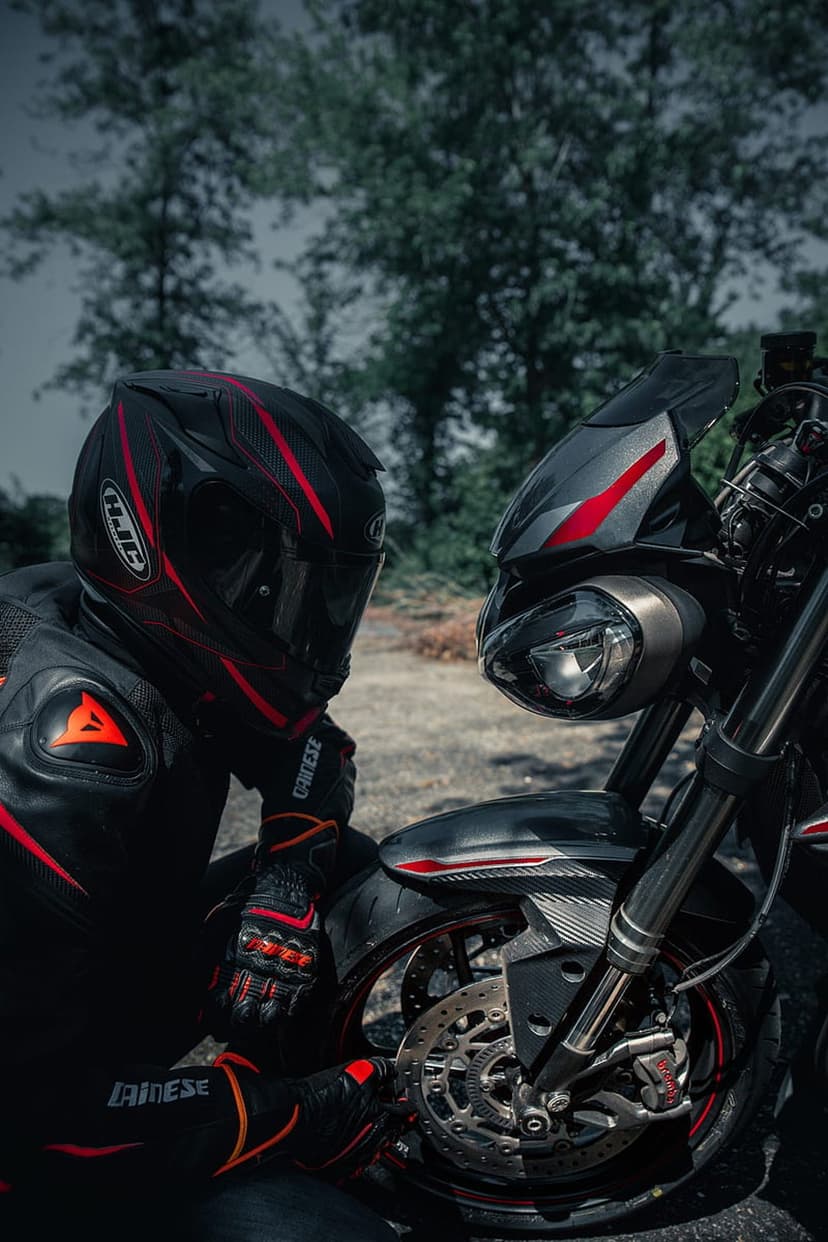 Motorcyclist in a red and black helmet stands by a sportbike with visible headlights against a blurred forest.