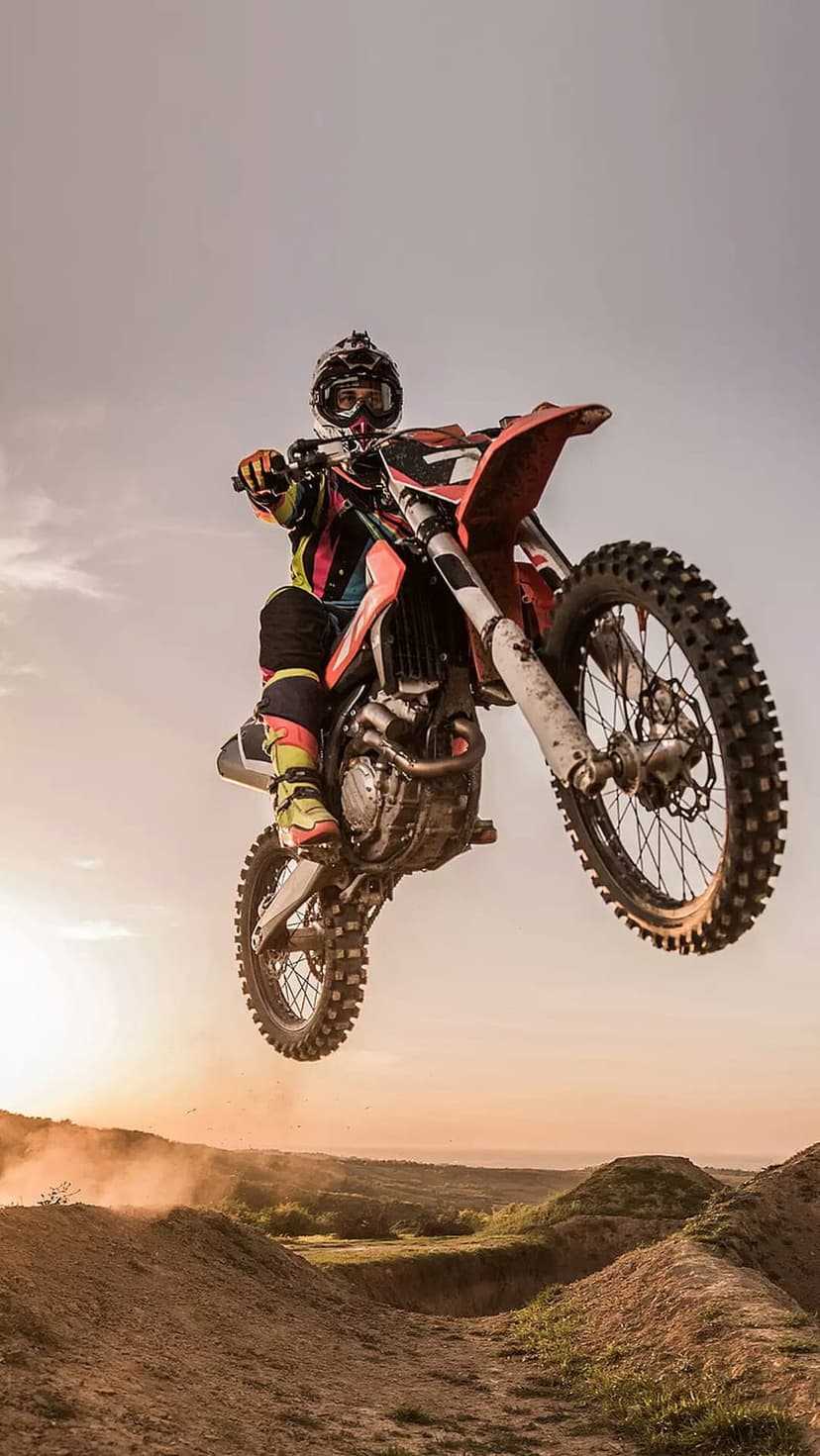 Motocross rider performing a high jump on a motorcycle against a dusty orange sunset sky during golden hour.