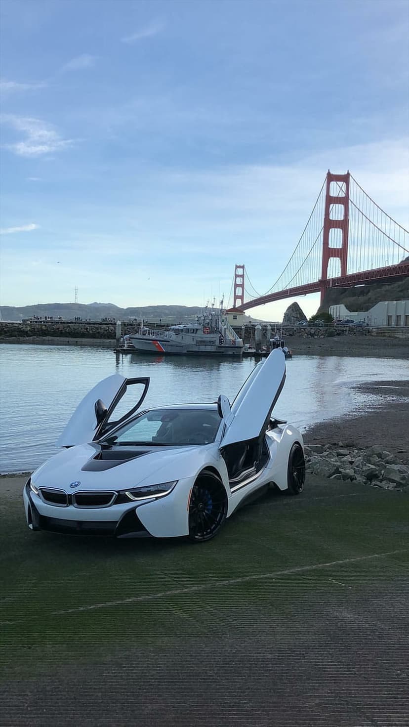 White BMW i8 sports car with scissor doors open parked by the Golden Gate Bridge and a Coast Guard ship.