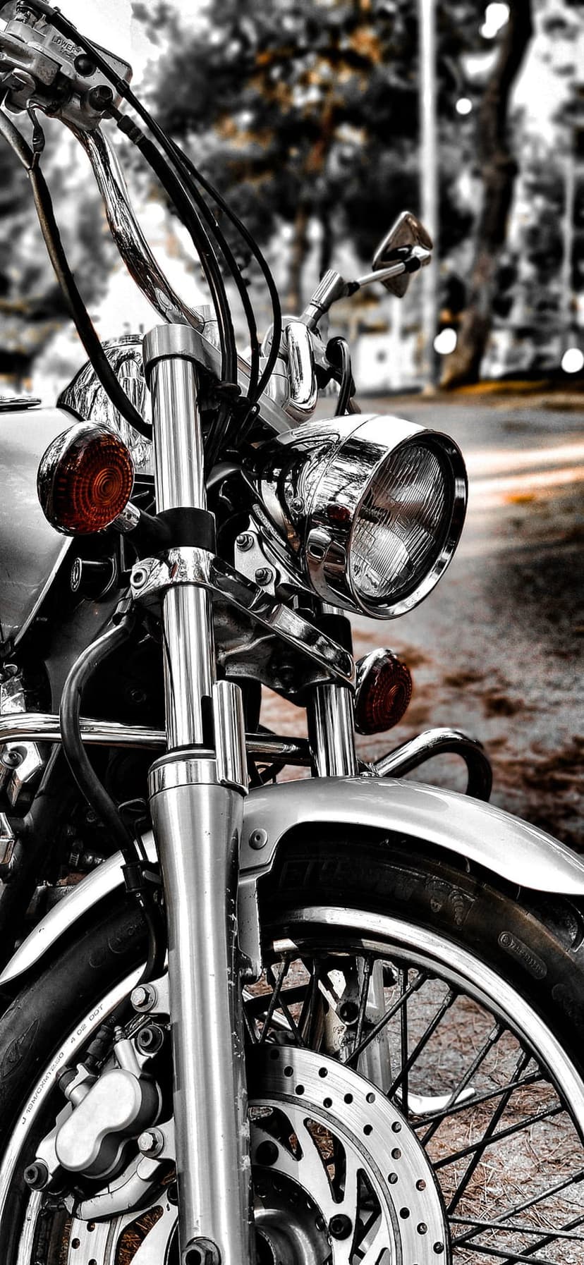 Chrome motorcycle headlight and spoked wheel with a red indicator light against a blurred outdoor background.