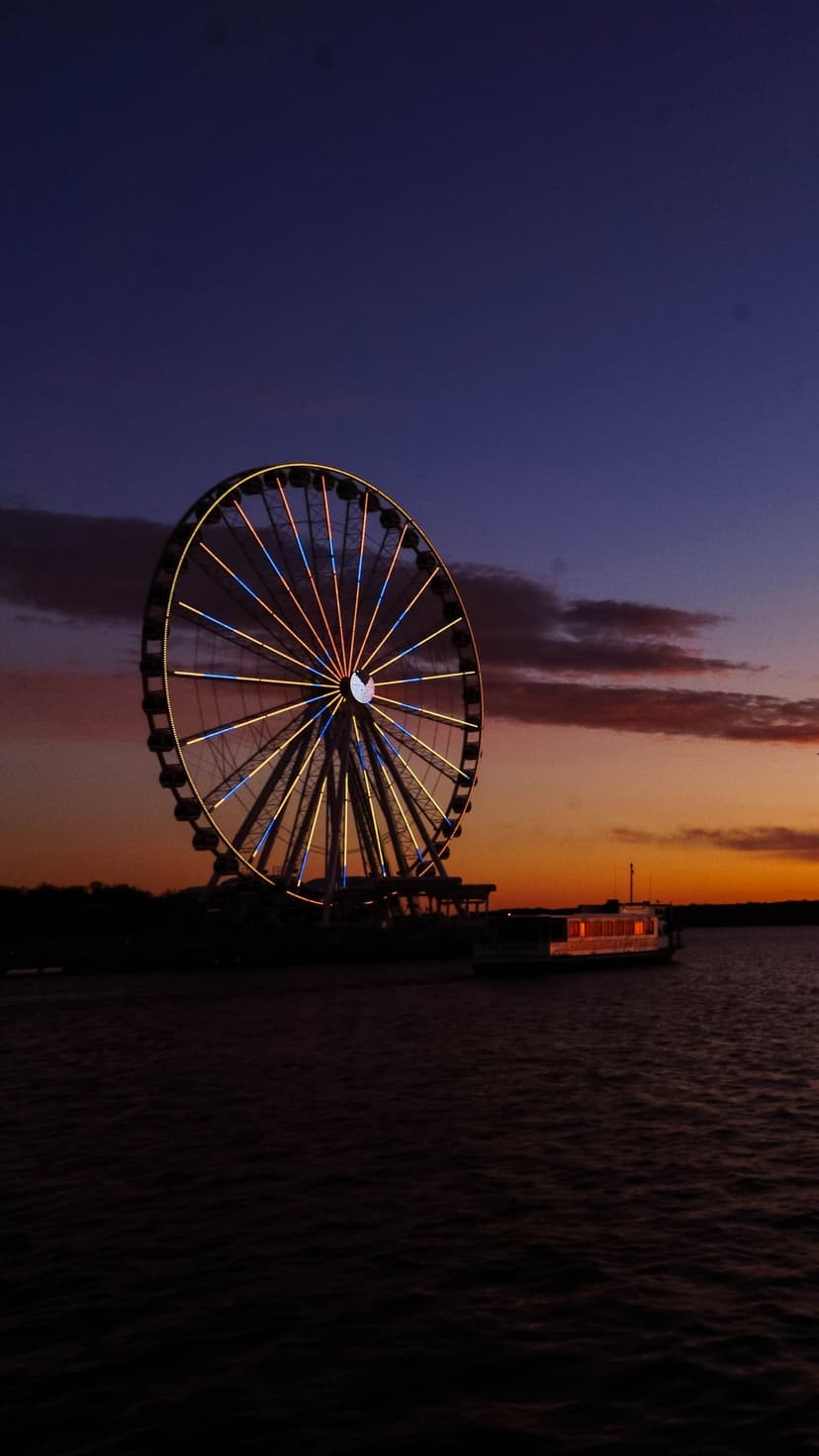 Ferris wheel with yellow and blue lights stands by a river under an orange and indigo sunset sky with a boat.