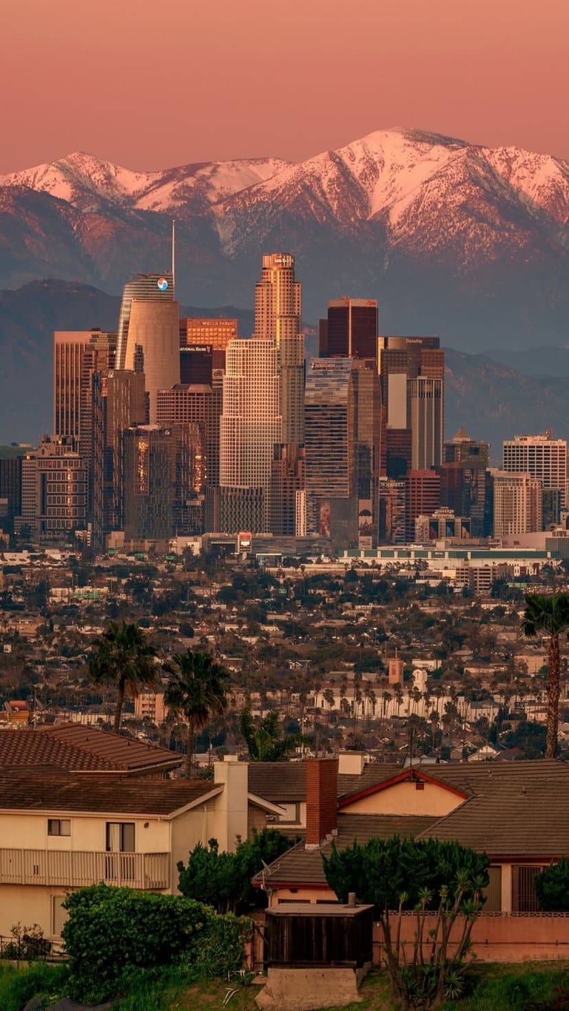 Los Angeles city buildings stretch toward snow-covered mountains under a vibrant pink and orange sunset sky.