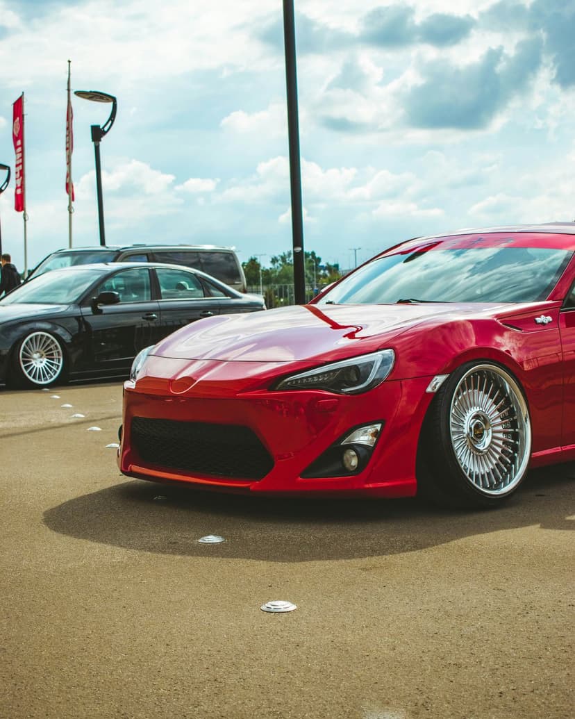 Red sports car with custom wheels and a lowered stance parked on asphalt under a cloudy sky at a car show.