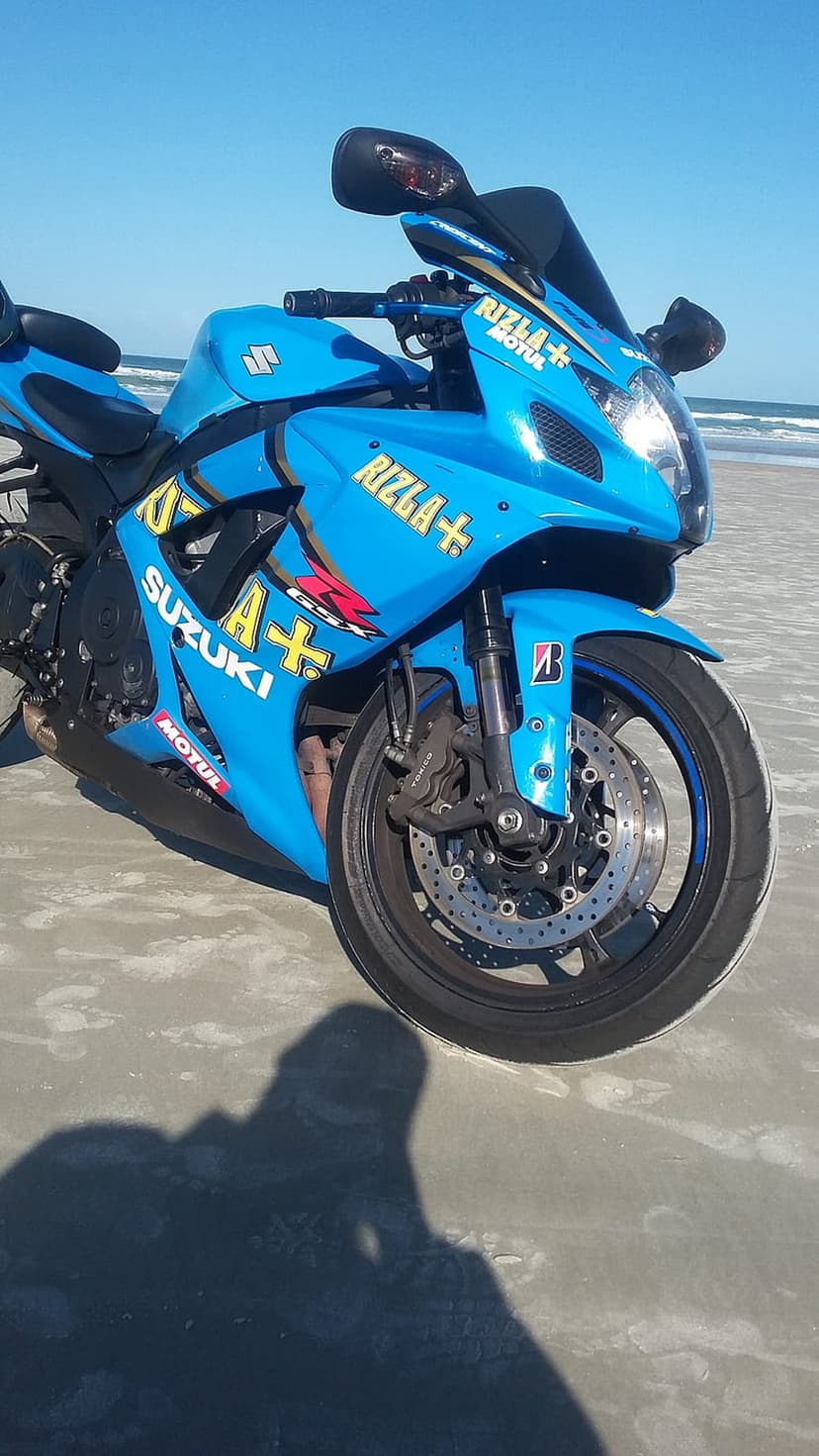Blue Suzuki sportbike with Rizla decals parked on sand with ocean waves and clear sky in the coastal background.